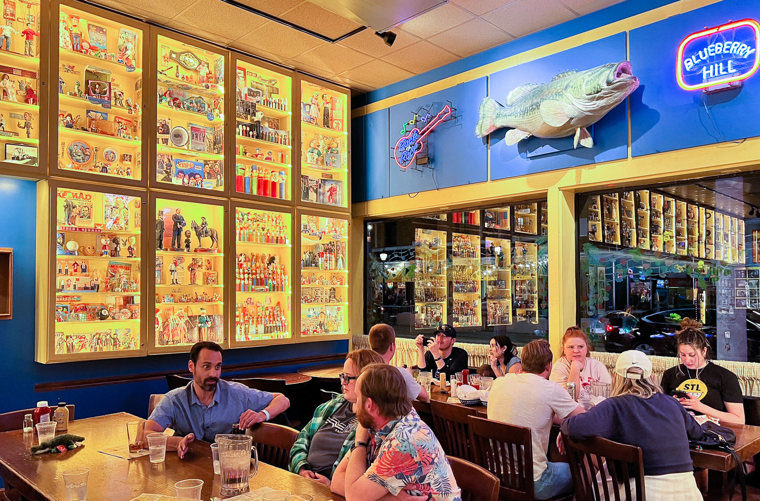 The Dart Room at Blueberry Hill. Two groups of people enjoy food and beer at two tables. There are neon signs and large colorful display cases of memorabilia around them.