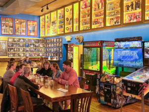 The Dart Room at Blueberry Hill. A group of people enjoy food and beer at a table. There are video games and large colorful display cases of memorabilia around them.