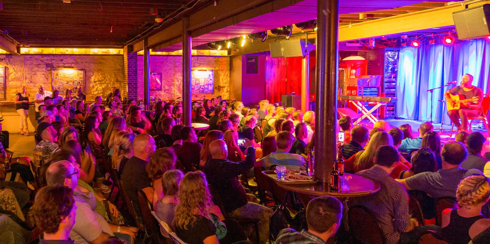 A large audience sits in a dimly lit music venue, facing a stage where a performer plays guitar and sings under colorful lights. The room is filled with people, tables, and a warm, vibrant atmosphere.