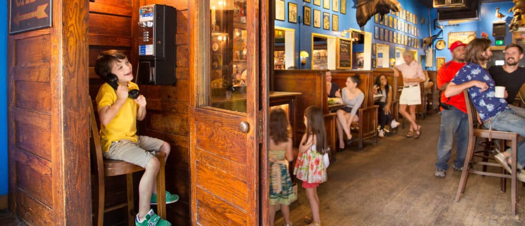 A young boy sits in a wooden phone booth using a payphone, while people of various ages talk and relax in a lively, rustic restaurant with blue walls and animal mounts.