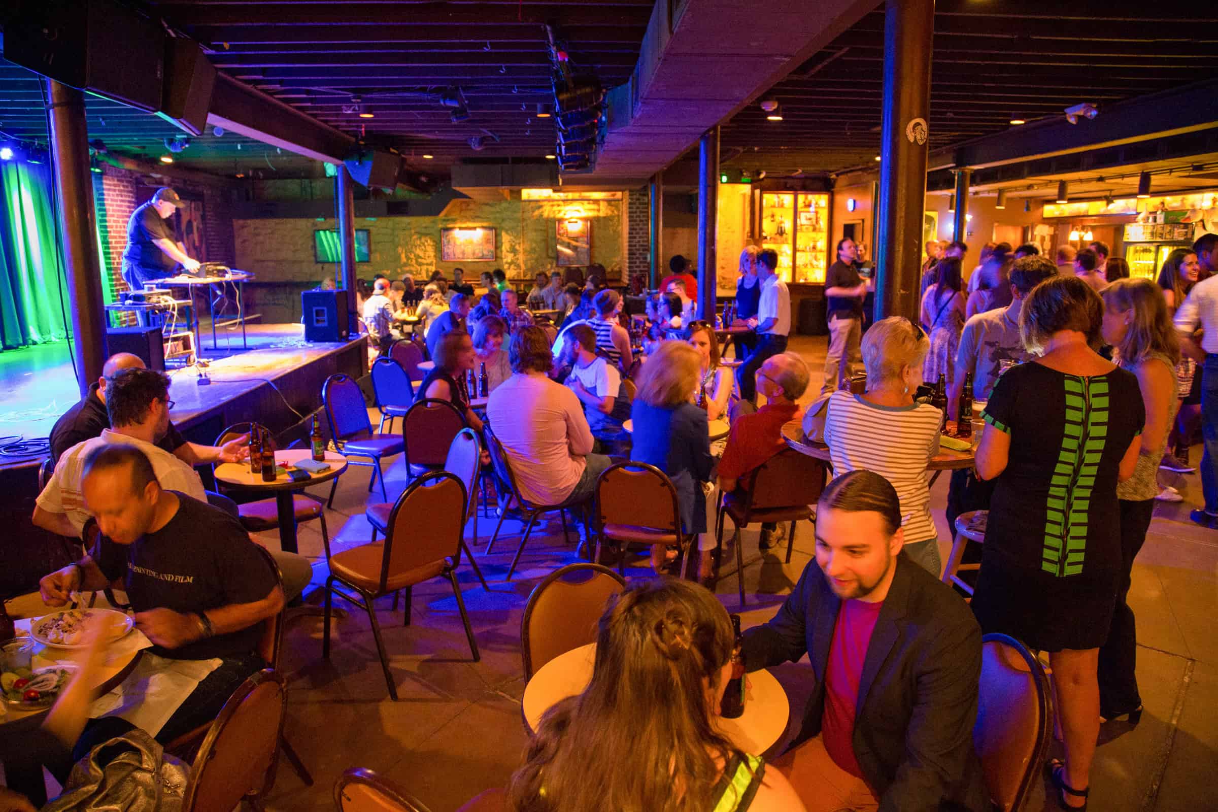 A lively indoor venue with people seated at tables, eating, drinking, and socializing. A performer is on stage with equipment under colorful lights. The atmosphere is vibrant and busy.