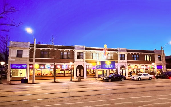 A brightly lit two-story building with neon signs and large windows, featuring parked cars in front; the sky is a vivid blue at dusk, and streetcar tracks run along the road.