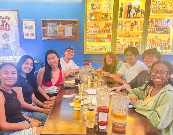 A group of eight young adults sit around a wooden table at a restaurant, smiling at the camera. There are drinks, condiments, and pitchers on the table, with colorful collectibles displayed on shelves in the background.