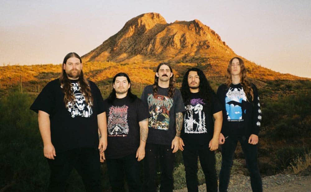 Five men with long hair stand outdoors in front of a rocky hill at sunset, wearing dark graphic T-shirts with band logos and jeans, posing together for a group photo.