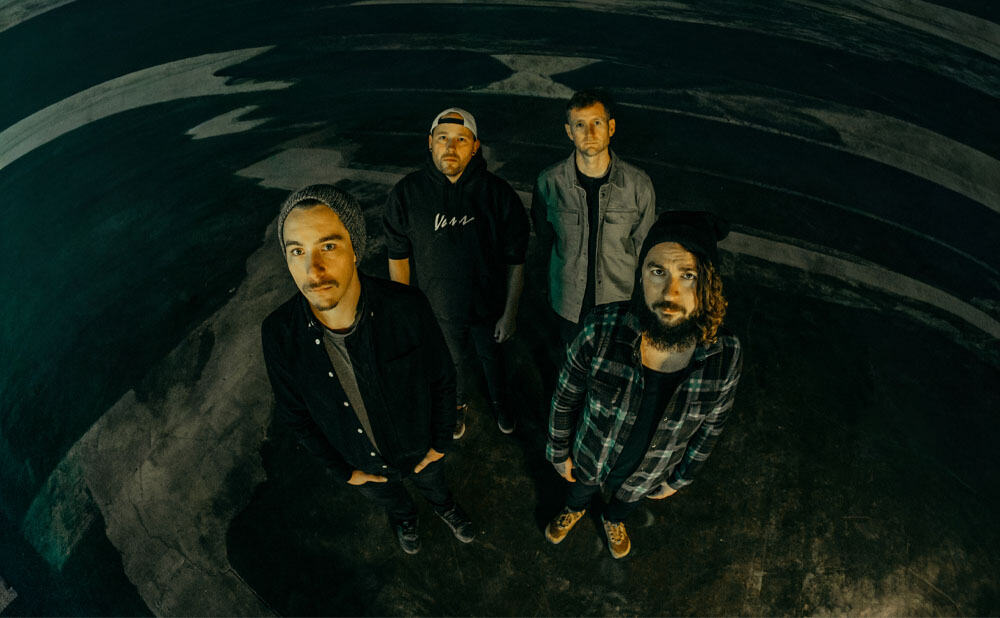 Four men stand in a wide, dimly lit space at Blueberry Hill Duck Room, looking up at the camera. They wear casual clothes—jackets, hats, flannel shirts—against a dark, abstract painted background.