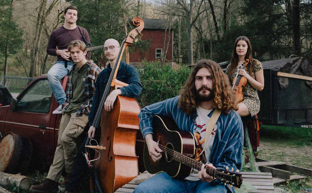 Five musicians pose outdoors with their instruments, including a guitar, upright bass, and fiddle. Surrounded by trees and rustic elements, they evoke the vibe of Cole Chaney at Blueberry Hill Duck Room. A red building and parked vehicle sit in the background.